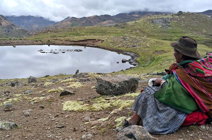 Mujer altoandina descansa junto a una laguna, con alpacas al fondo