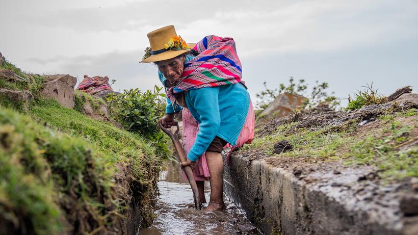 Mujer andina limpiando un canal de riego con su herramienta
